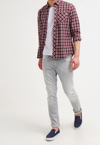 Mies, joka kävelee punamustassa ruudullisessa paidassa valkoisen t-paidan päällä, vaaleanharmaissa farkuissa ja tummansinisissä liukuvaatteissa, valkoista taustaa vasten.