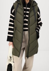 Osoba nosząca oliwkowo-zieloną pikowaną kamizelkę na czarnym i beżowo-paskowanym swetrze, beżowe spodnie, trzyma kremową i czarną teksturowaną torbę na ramię.