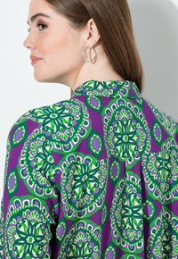 Violetti paita, jossa on monimutkaiset vihreät ja valkoiset kukkakuviot, kaulus ja kangassa on hieno kiilto. Hopeiset rengaskorvakorut.