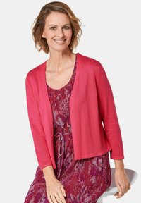 Roze cardigan met een ontspannen pasvorm, open voorkant en lange mouwen, gecombineerd met een jurk met bloemmotief in paarse en rode tinten.