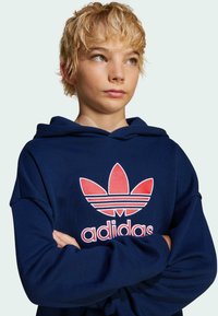 Sweat à capuche bleu marine en tissu doux, présentant un grand logo Adidas rouge et un design de trois bandes à l'avant.