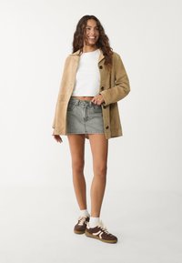 Beige mockaljacka med öppen krage och mörka knappar, bärs över en vit kort topp och en jeanskjol, ihopparat med bruna sneakers.