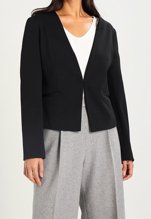 Vrouw die een zwarte, op maat gemaakte blazer draagt over een witte top en grijze wijde broekspijpen tegen een effen lichte achtergrond.