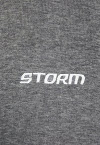 Harmaa puuvillakangas, jossa on valkoisella brodeerattu teksti "STORM". Kankaan pinta näyttää pehmeältä, ja siinä on hienovarainen heather-kuvio.