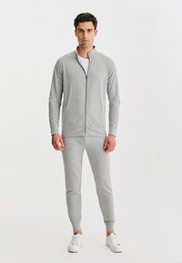 Szara bluza na zamek z bocznymi kieszeniami, w komplecie z dopasowanymi spodniami dresowymi zwężanymi ku dołowi. Gładka tkanina i ściągacze na mankietach, noszone z białymi trampkami.