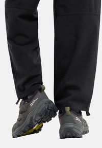 Dunkelgraue Wanderschuhe mit robusten strukturierten Sohlen, die gelbe Akzente und ein gesichertes Schnürsystem aufweisen, kombiniert mit schwarzen Hosen.