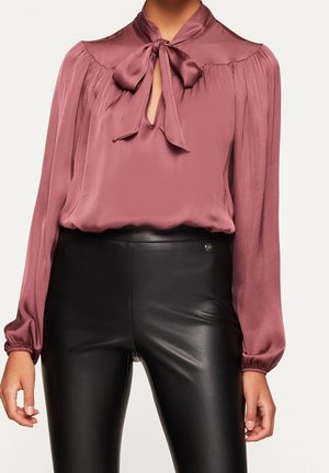 Vrouw draagt een mauve satijnen blouse met een gestrikte strik bij de hals en lange pofmouwen, gecombineerd met een hooggesneden zwarte leren broek.