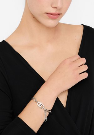 Silberarmband mit verbundenen Ketten, einem mittig angebrachten ovalen Anhänger und einem dekorativen Schlosselement, getragen an einem nackten Handgelenk vor schwarzem Hintergrund.