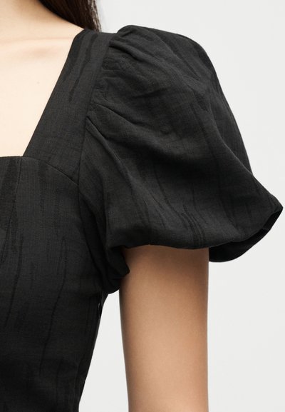 Μαύρο φόρεμα με κοντά φουσκωτά μανίκια και διακριτή κάθετη υφή στον τετράγωνο λαιμόκοψη, που φοριέται από ένα άτομο με μακριά σκούρα μαλλιά.