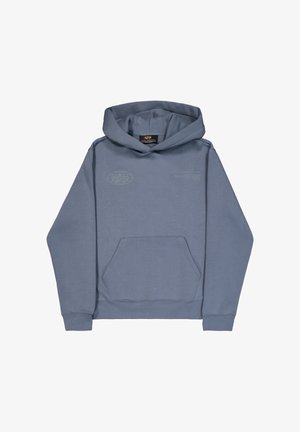 Osoba s pletenicama, odjevena u svijetloplavu hoodicu Alpha Industries i crne cargo hlače, okrenuta leđima na jednobojnoj pozadini.