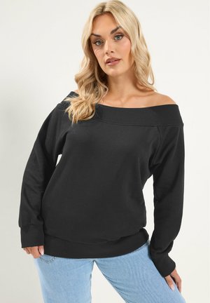 Lyshåret kvinde iført en off-shoulder sort langærmet top og lyseblå jeans mod en lys baggrund.