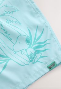 Lichtblauwe zwemshorts met een afbeelding van een cartoonfiguur en bladeren. Label met de tekst "SNOOPY." Gladde textuur met eenvoudige stiksels.