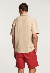 Camisa beige de manga corta con un corte relajado y tejido texturizado, combinada con pantalones cortos rojos, vista desde la parte posterior.