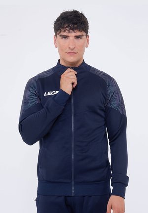 Uomo con capelli scuri e ricci che indossa una giacca blu navy con zip e motivi geometrici sulle spalle, tiene la zip vicino al colletto.