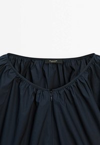 Mörknavy blus med vid halsringning och rynkade detaljer. Har en central dragkedjestängning och en etikett som säger "Massimo Dutti." Slät textur.