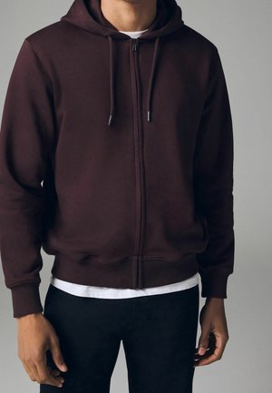 Homme portant un sweat à capuche zippé bordeaux foncé avec des cordons, un t-shirt blanc et un pantalon noir, debout devant un fond gris uni.