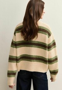 Cremefarbener Pullover mit grünen und braunen Streifen, oversized Schnitt, gerippter Saum und weicher Textur, kombiniert mit dunklen Jeans.