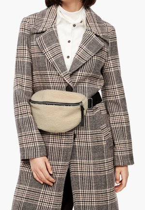 Ce sac banane en polaire beige avec une fermeture éclair noire est porté par-dessus un manteau à carreaux gris aux grands revers, avec une chemise blanche en dessous.
