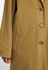 Abrigo color caqui con una tela texturizada, que presenta un frente abotonado, un bolsillo en el pecho y costuras visibles a lo largo de los bordes.