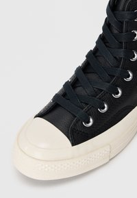 Černé kožené vysoké tenisky s bílým gumovým špičkou a texturovanou podešví; obsahují tmavé tkaničky a stříbrné dírky na tkaničky.