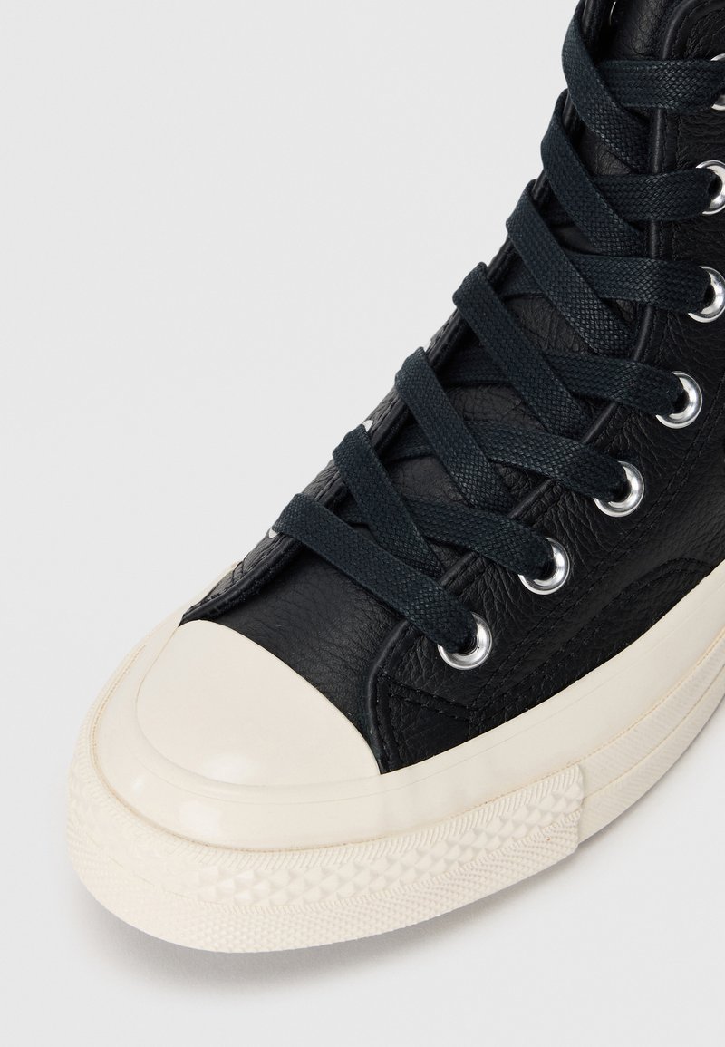 Sneaker alto in pelle nera con punta in gomma bianca e suola testurizzata; presenta lacci scuri e occhielli in argento.