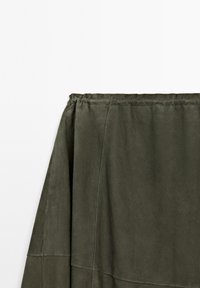 Oliven grøn imiteret ruskind nederdel med samlet talje, som har en A-linjeform og synlige syninger. Glat tekstur med subtil glans.