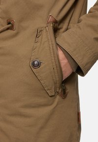 Veste kaki avec une fermeture éclair et un bouton. Comprend des accents en cuir, un tissu texturé et une poche latérale pour le rangement.