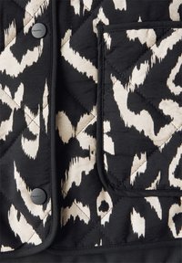 Gestepptes Stoff in Schwarz und Creme mit abstrakten Mustern, ausgestattet mit einer Brusttasche und schwarzen Knöpfen. Glatte Textur und saubere Nähte.