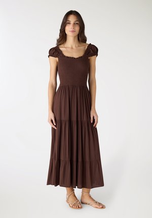 Donna che indossa un abito lungo marrone a maniche corte a balze con corpetto arricciato, in piedi contro uno sfondo bianco semplice.