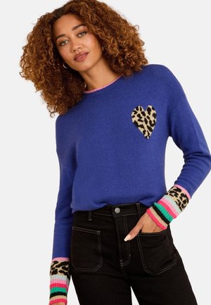 Vrouw met krullend haar die een blauw trui draagt met een luipaardprint hart en kleurrijke gestreepte manchetten, gecombineerd met zwarte hooggetailleerde spijkerbroek.