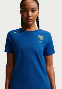 Blå kortärmad t-shirt i bomull med en gul Nike-swoosh och en gul Barcelona-logotyp på vänstra bröstet.