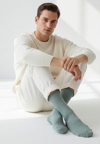Hombre con cabello corto y castaño vestido con un jersey y pantalones crema, sentado con las piernas cruzadas, usando calcetines verde azulado con lunares blancos en una habitación luminosa.
