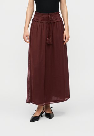 Femme portant une jupe bordeaux taille haute, longueur cheville, avec une taille à cordon et des pompons, associée à des talons noirs à bouts pointus.