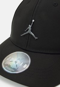 Svart caps laget av teksturert stoff med en blank sølv Jumpman-logo og et holografisk sirkulært merke med "JORDAN" trykket.