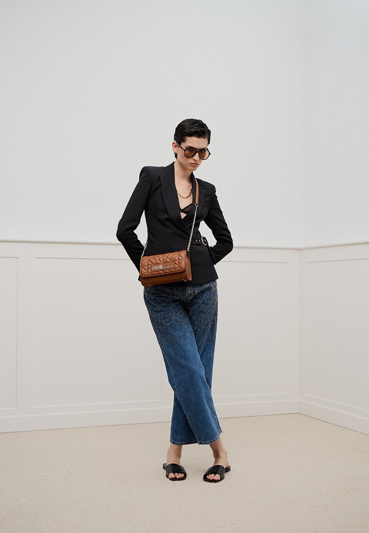 Blazer noir ajusté sur un haut noir, associé à un jean bleu ample. Sac bandoulière matelassé marron, lunettes de soleil surdimensionnées et sandales noires.