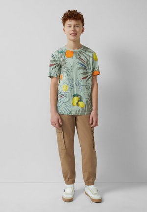 T-shirt vert à manches courtes avec un imprimé sur le thème des fruits, pantalon cargo beige et baskets blanches avec une semelle marron. Design simple et décontracté.