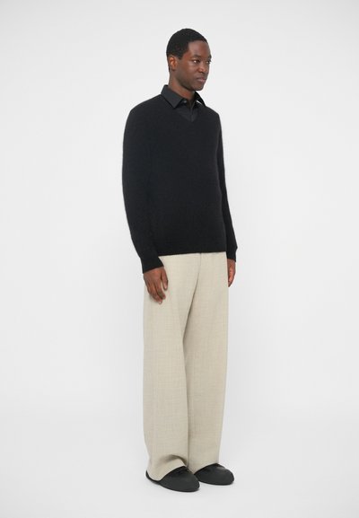 Maglioncino in misto lana nero, a scollo a v, indossato sopra una camicia nera con colletto. I pantaloni beige chiaro a gamba larga e le scarpe nere senza lacci completano l'outfit.