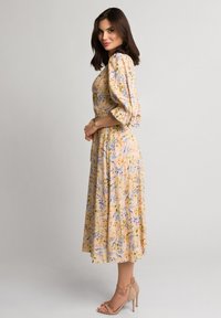 Kwiatowa sukienka midi w jasnym żółtym kolorze z niebieskimi i pomarańczowymi wzorami, z bufiastymi rękawami, talią podkreśloną wcięciem oraz zwiewnym dołem, w zestawieniu z cielistymi sandałami na obcasie.
