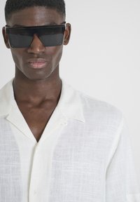 Camisa de lino blanca de manga corta con un cuello relajado, detalle de botones y un patrón sutilmente texturizado. Unas gafas de sol negras de gran tamaño la complementan.