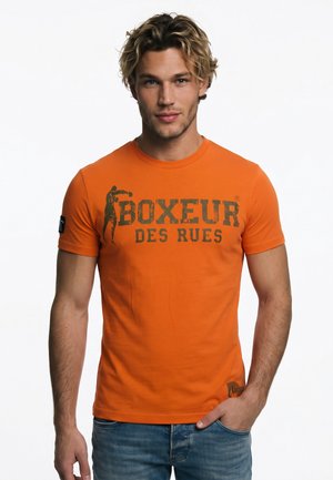 Joven con cabello rubio ondulado que lleva una camiseta naranja con el texto "BOXEUR DES RUES" y un gráfico de boxeador, combinado con vaqueros azules.