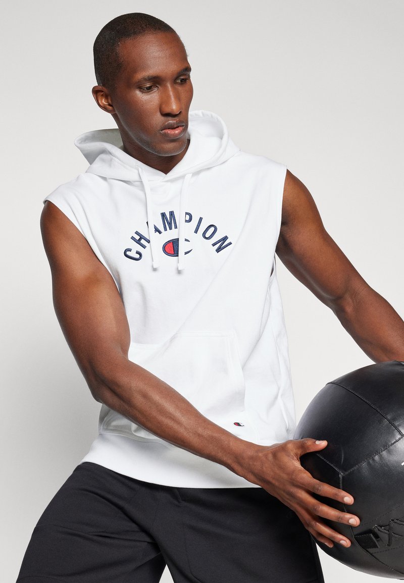 Weißer ärmelloser Hoodie mit "CHAMPION" in Marineblau bedruckt, ausgestattet mit einer Fronttasche und einer Kapuze mit Kordelzug, kombiniert mit schwarzen Sportshorts.