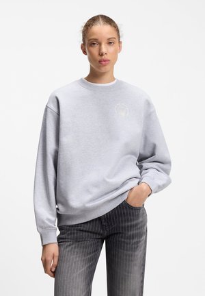 Junge Frau mit zurückgebundenem Haar, die einen hellgrauen Pullover und dunkelgraue Jeans mit Nadelstreifen trägt, mit einer Hand in der Tasche.