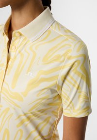Żółta koszulka polo z falistym, abstrakcyjnym wzorem, krótkimi rękawami, dwuguzikową plisą oraz kontrastującym białym kołnierzem z dwoma paskami.