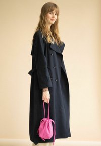 Laivyno spalvos dvigubai užsegamas paltas su dideliais apykaklėmis ir kišenėmis, derinamas su mažais rožiniais maišeliais su raišteliais. Modelis stovi prieš neutralų foną.