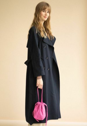 Abrigo azul marino de doble botonadura con grandes solapas y bolsillos, combinado con una pequeña bolsa rosa con cordón. El modelo se encuentra de pie frente a un fondo neutral.