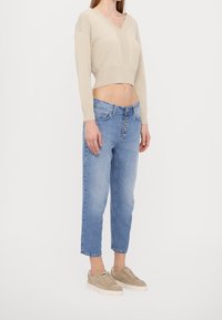 Mujer vestida con un suéter corto de pico beige, jeans azul claro de tiro alto con botones, y zapatillas beige, de pie de lado sobre un fondo blanco.