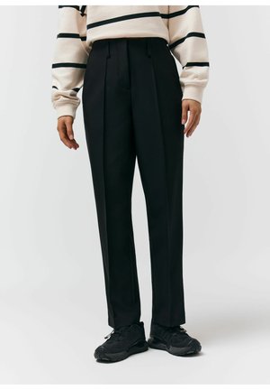 Osoba ubrana w czarne, dopasowane spodnie z plisami z przodu oraz czarne trampki, w połączeniu z kremowym swetrem w czarne paski.