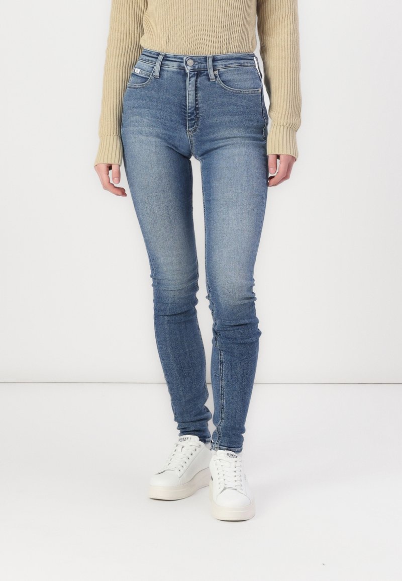 Blauwe jeans met hoge taille in een vervaagde wassing en een slanke pasvorm. Voorzien van lussen voor een riem, steekzakken aan de voorkant en een gladde textuur. Gecombineerd met witte sneakers.
