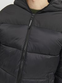 Μαύρο επενδυμένο μπουφάν με κουκούλα, διαθέτει κλείσιμο με φερμουάρ και σχέδιο τσάκισης. Το υλικό φαίνεται λείο και ελαφρύ.