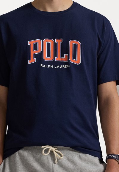 Jūras zilas kokvilnas T-krekls ar "POLO" oranžā un baltā krāsā, zem kura baltā krāsā ir "RALPH LAUREN". Īsas piedurknes un apaļa kakla izgriezuma.